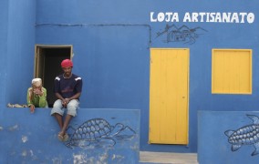 Two people sit on a wall in front of a blue house with yellow doors